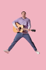 Young happy man with guitar jumping on pink background
