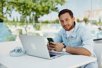 Young man working remotely on laptop, smiling while using smartphone at outdoor cafe