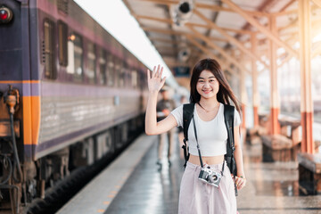 Smiling young woman with backpack and camera waving at a train station platform, ready for travel and adventure.