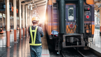 A worker in a safety vest and helmet stands on a train platform, facing a train during sunset,...