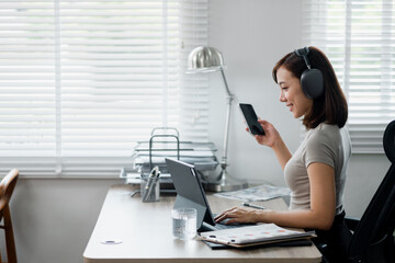 A young woman multitasking in a modern home office, using a laptop and smartphone while wearing headphones.