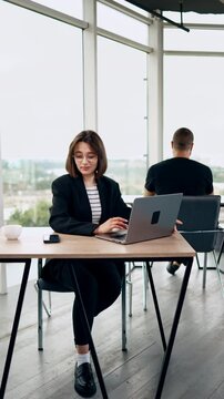 People work at desks in the spacious office. Woman in glasses works on laptop distracting on her smartphone. Vertical video.