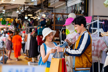 Happy Asian man and woman couple enjoy and fun outdoor lifestyle travel in the city shopping and buying fashion clothes together at street market in Bangkok, Thailand on summer holiday vacation.