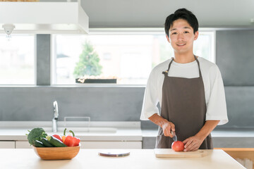 キッチンで料理・家事をする若いアジア人男性・主夫・夫（包丁・切る・野菜・トマト）  © buritora