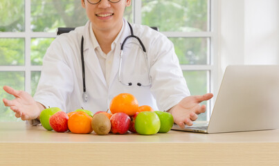 Fototapeta premium Smiling male nutritionist doctor in white coat sitting at clinic office desk with laptop and gesturing of colorful of apples, oranges and kiwis fruits. Nutrition and healthy eating concept