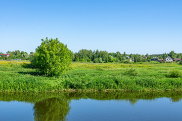 Summer countryside landscape with village and river under blue sky. Rural river with green meadow peaceful landscape.