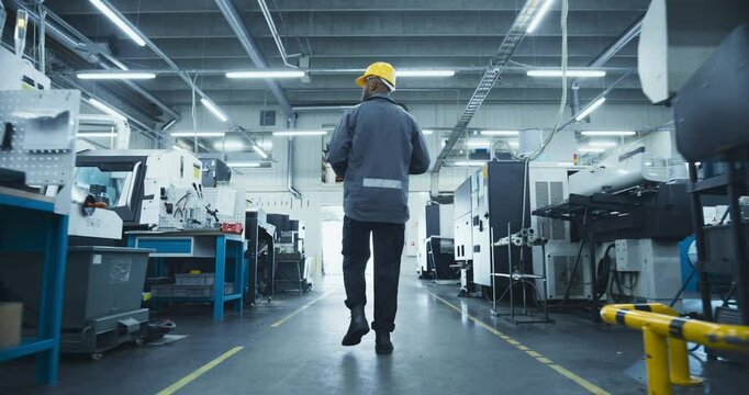 Black Male Worker in a Yellow Hard Hat Walking Through a Modern, Well-lit Factory, Inspecting Advanced Machinery, Checking Equipment, Working in Production Industry. Footage From the Back