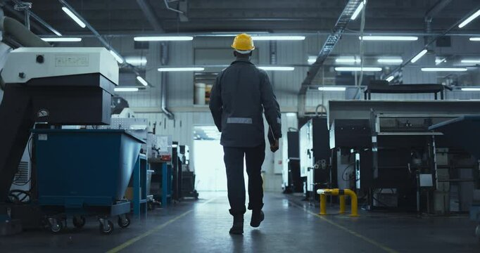 Young Specialist Walking in a Metal Components Manufacturing Facility at a Factory. Industrial Engineer Wearing Uniform and a Hard Hat for Safety. Follow Footage From the Back