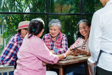 Elderly Asian women sitting at a wooden table in a bright, sunlit cafe, laughing and enjoying their time together. Waiter standing by, ready to take their order. Atmosphere is warm, friendly,