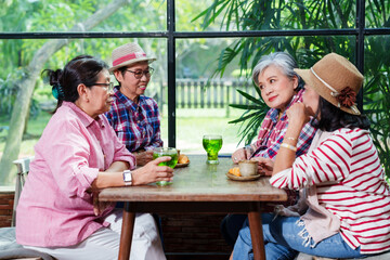 Group of elderly Asian women sitting at a wooden table in a bright cafe, having a friendly conversation and enjoying drinks. Casual attire, one woman wearing a hat.