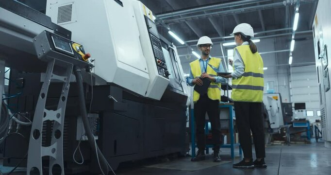 Two Industrial Engineers at Work. Caucasian Female with a Laptop and a Middle Eastern Male Monitor a CNC Machine in a High Tech Metal Factory, Ensuring Precision and Efficiency in Automation Processes