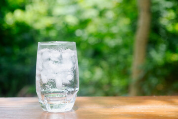 a glass of cold water with ice on wooden table in garden background.