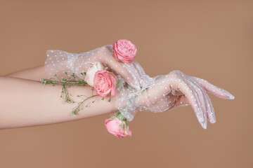 A pair of hands wearing white dotted lace gloves are adorned with pink roses and greenery. The image is shot against a soft brown background - Powered by Adobe