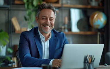 A smiling middle-aged man in a blue suit sits at a desk in an office, working on a laptop