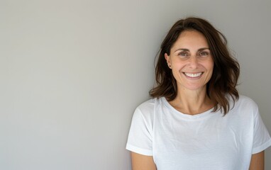 Fototapeta premium A brunette woman with a bright smile poses against a light grey wall, wearing a white t-shirt