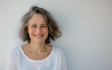 A woman with short, grey hair is smiling at the camera. She is wearing a white t-shirt and standing in front of a plain white wall