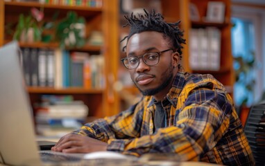 A young African American man with dreadlocks, wearing glasses and a plaid shirt, works on a laptop computer at a desk in a home office
