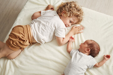 Charming little brother and sister lying together on white sheet on bed wavy haired blond older...
