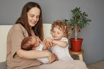 Joyful and affectionate family. Smiling Caucasian mother engaging with her newborn child and her older curly blonde-haired daughter relaxing on the sofa in living room together