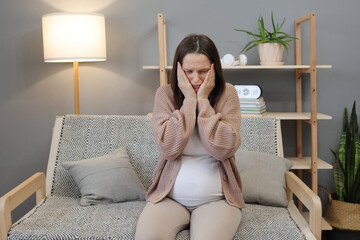 Sad upset pregnant woman wearing casual clothes sitting comfortably on sofa at home expressing...