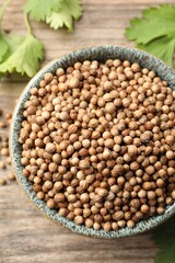 Dried coriander seeds in bowl and green leaves on wooden table, flat lay