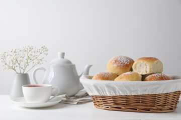Delicious dough balls with powdered sugar in basket, tea, teapot and gypsophila flowers on white wooden table