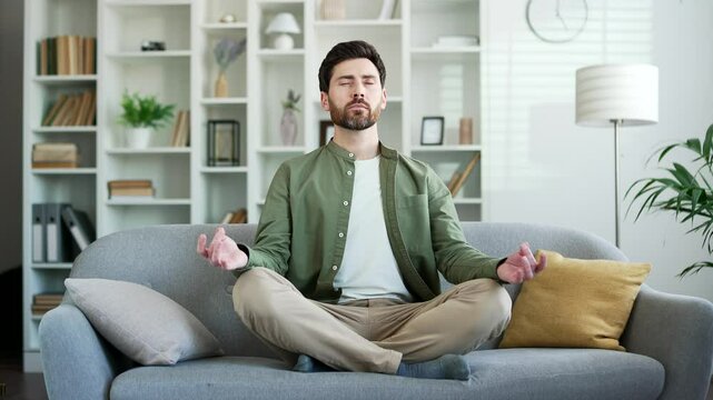 Handsome man meditating with eyes closed while sitting on sofa in living room at home. Calm happy bearded male relaxing in lotus position, practicing yoga on comfortable couch breathing deeply