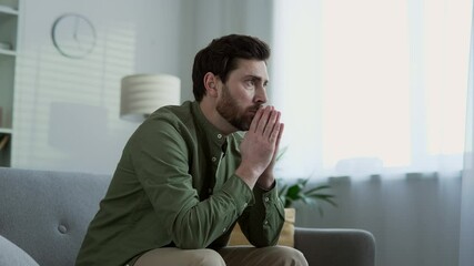 Sad male suffering from depression and mental pain sitting on sofa in living room at home. Lonely bearded man thinking about his problems. He is unhappy, disappointed, has a bad mood, feels anxious