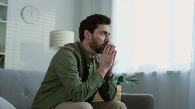 Sad male suffering from depression and mental pain sitting on sofa in living room at home. Lonely bearded man thinking about his problems. He is unhappy, disappointed, has a bad mood, feels anxious