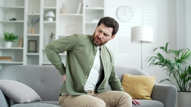 Tired man suffering from back pain while sitting on sofa in living room at home. Upset mature bearded male holds his lower back with his hands, massages painful muscles, has sciatica or a sick spine