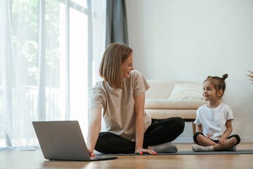 Using laptop. Young woman with little girl are doing yoga at home