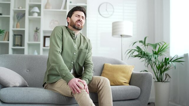 Handsome man suffers from painful severe knee joint pain while sitting on sofa in living room at home. Upset bearded male massages leg muscles, could not walk, illness, cramps, rheumatism