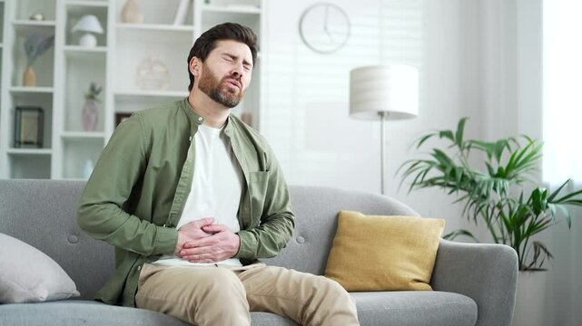 Sick man feels stomach pain while sitting on sofa in living room at home. Upset handsome bearded male has heartburn, gastritis, bloating or poisoning. Male suffers from spasms and constipation