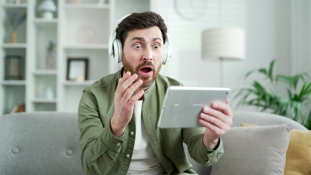 Excited handsome man in wireless headphones watching sports match competition on digital tablet sitting on sofa in living room at home. Happy bearded male emotionally cheering for favorite team