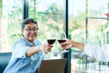 Elderly Asian woman in a blue denim shirt toasting with another person in a modern restaurant. Both holding glasses of wine, surrounded by natural light, large windows, green plants.