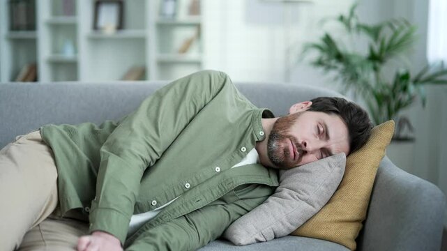 Exhausted man tired after hard working day lies down on sofa in living room at home. Overworked overdriven male wants to sleep, falls off his feet on couch, puts his head on pillow and closes his eyes
