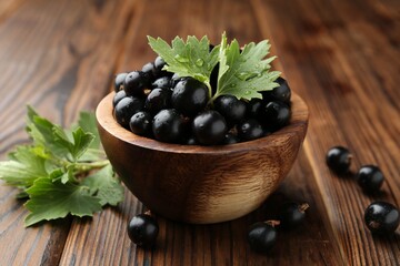 Ripe black currants and leaves in bowl on wooden table, closeup