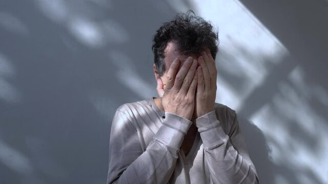 A middle-aged man with disheveled hair holds his face with both hands, expressing deep concern or stress. The light and shadow play on the background adds a dramatic effect. Copy space.