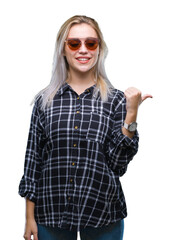 Young blonde woman wearing sunglasses over isolated background smiling with happy face looking and pointing to the side with thumb up.