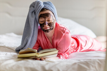 Woman with black face mask and towel on head reading book on bed