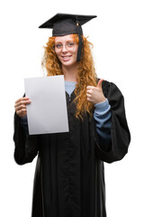 Young redhead woman wearing graduate uniform holding degree happy with big smile doing ok sign, thumb up with fingers, excellent sign