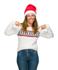 Middle age adult woman wearing winter sweater and chrismat hat over isolated background looking confident with smile on face, pointing oneself with fingers proud and happy.