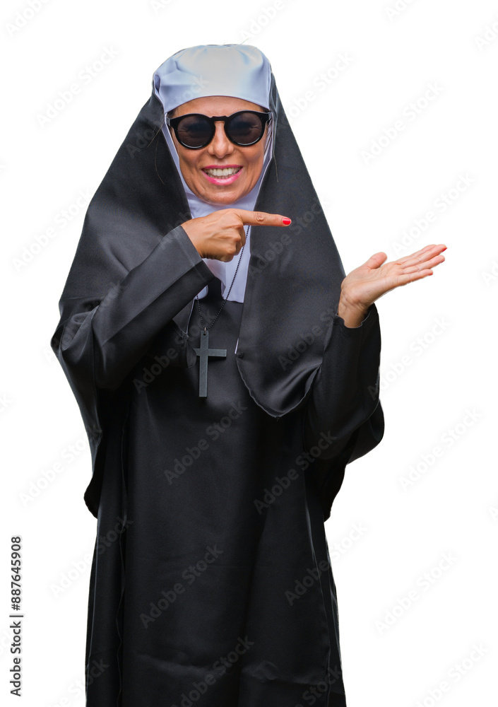 Wall mural Middle age senior catholic nun woman wearing sunglasses over isolated background amazed and smiling to the camera while presenting with hand and pointing with finger.