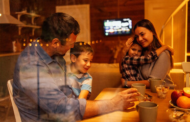 A young married couple in casual clothes with two cute smiling children are having dinner in the kitchen. Mom hugs her daughter. Dad holds his son in his arms.
