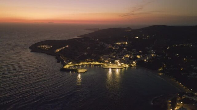A panoramic view of Stavros, Donousa, Greece, captured at dusk. The coastline is illuminated by streetlights, with the remaining landscape bathed in the soft glow of twilight.