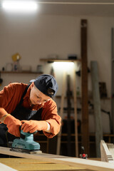 A female carpenter sands a wooden board using a sander while working in her carpentry workshop