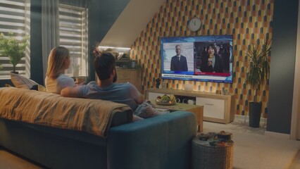 Caucasian couple watches news broadcast about United States Election Day on TV screen, talks and drinks tea sitting on couch at home. Political channel with breaking news with displayed on TV display.