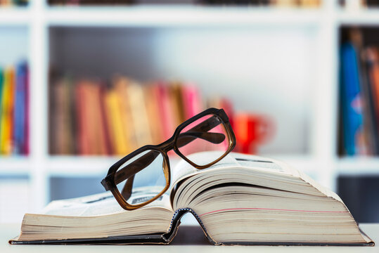 Reading glasses lying on opened book with blurred shelfbook of library in background