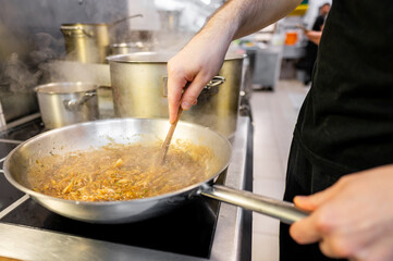 Close-up of a chef’s hand stirring a sizzling pan of food on a stove in a professional kitchen