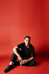 A stylish Asian man in black attire sits gracefully on the floor against a striking red background.